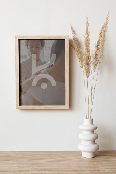 Cadre en bois avec une illustration abstraite et un vase blanc contenant des herbes sèches.